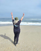 Person on a beach with arms raised holding up peace signs, facing the ocean and wearing a humpback whale design on the back of an organic t-shirt.