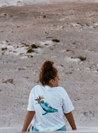 A person viewed from behind wearing a white organic t-shirt with a marine life design, standing in a natural, outdoor setting.