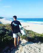 Surfer holding a surfboard with the beach in the background, wearing a black organic t-shirt with a whale design on the back.